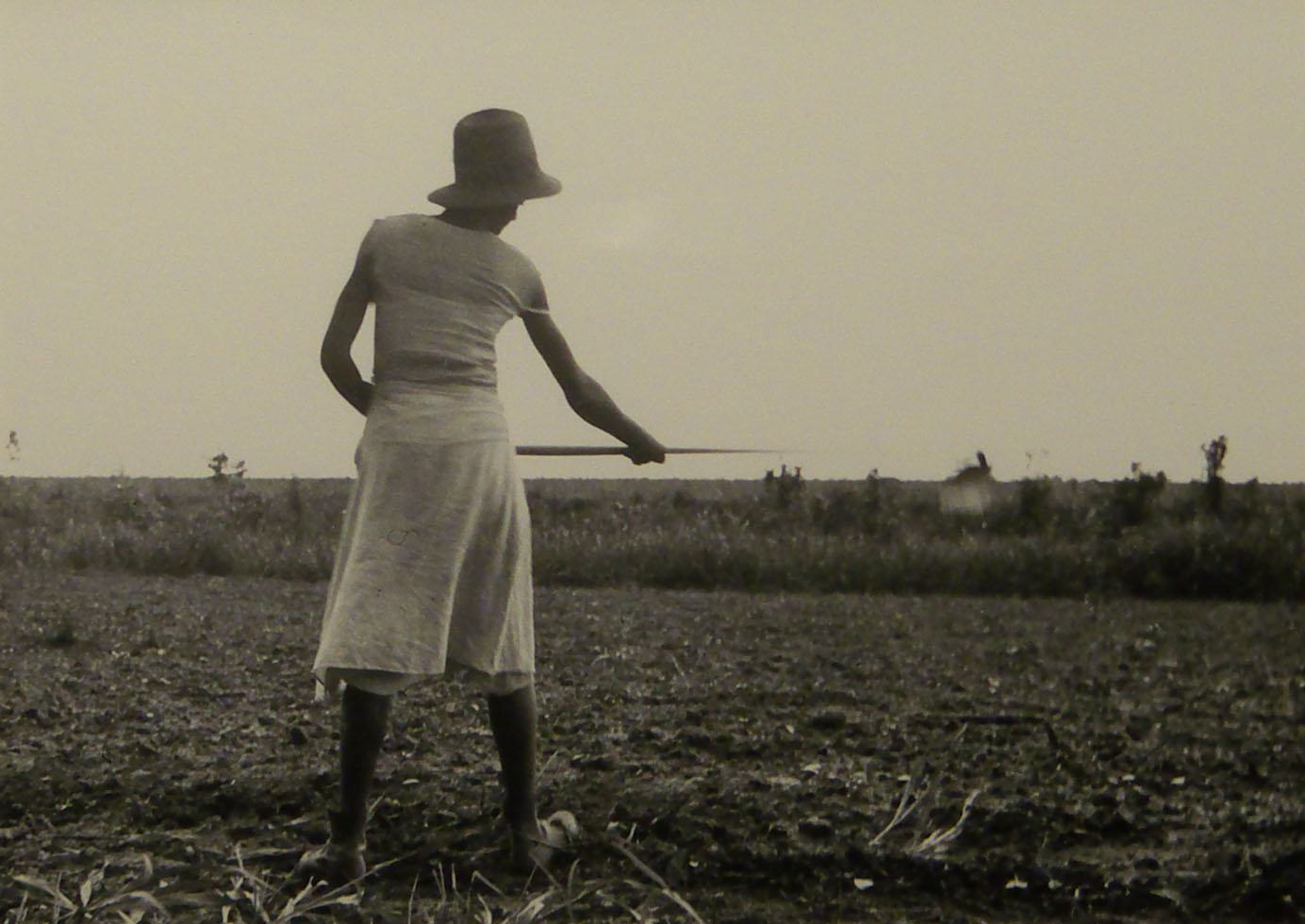 A woman in a dress wearing a hat holding a stick in a field.