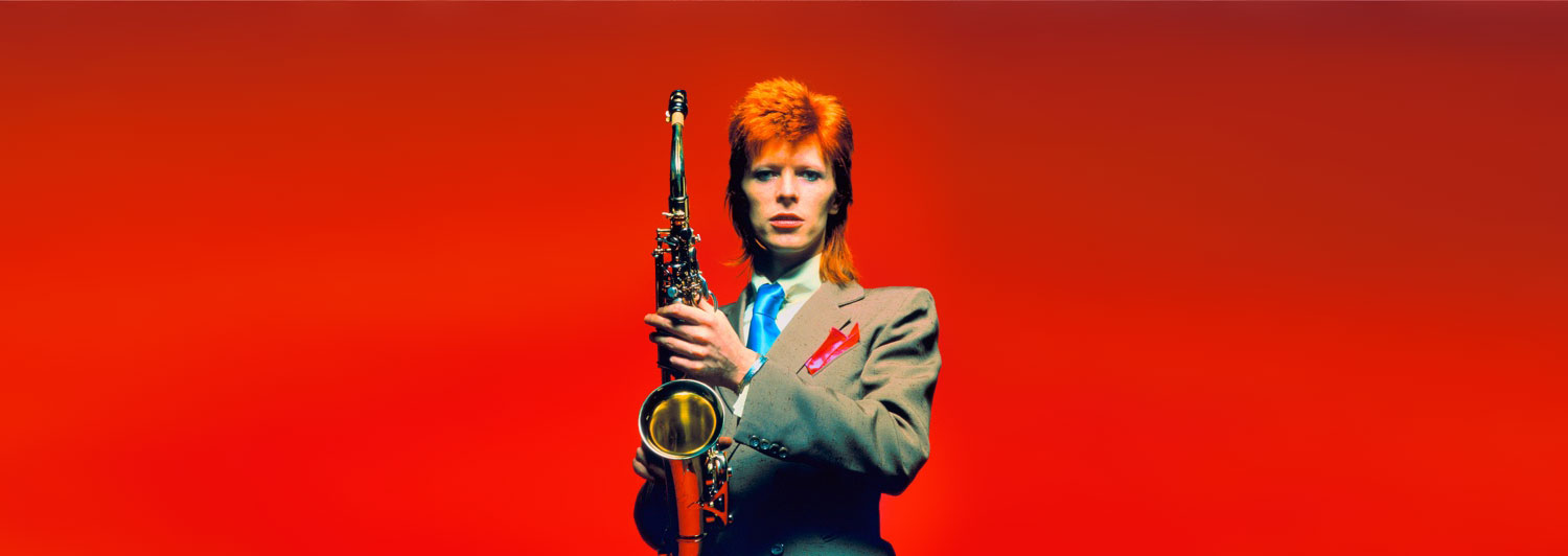 David Bowie in a suit holding a saxophone against a red background.