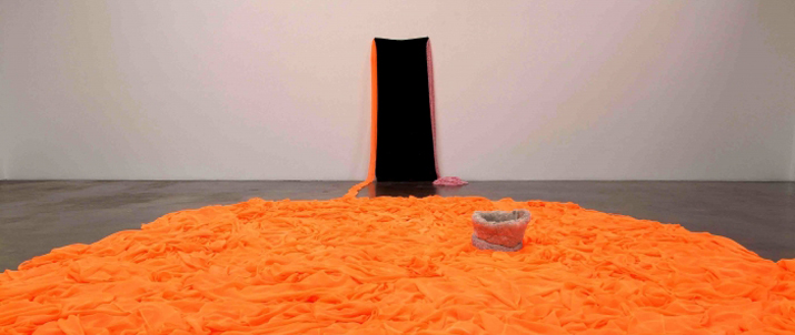 Orange tufted fabric is on the floor. There is a black piece of fabric on the wall with two streams of fabric leading to the pooled orange.