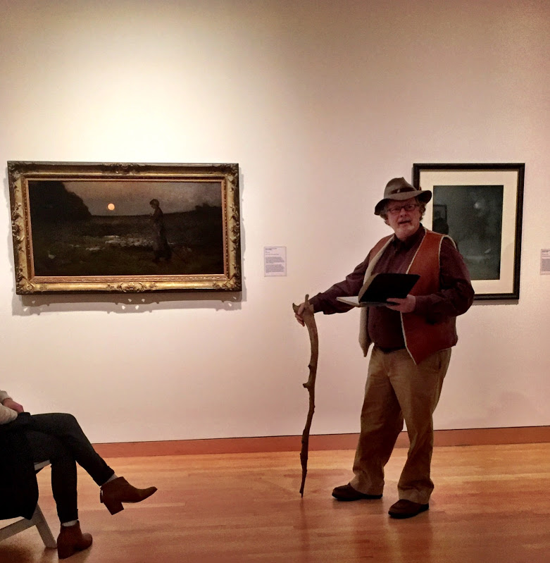 A man stands in front of artwork with a large walking stick and an open book.