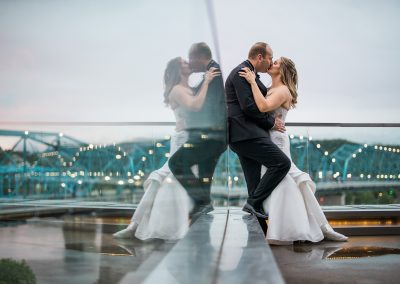 A bride and groom kiss on the balcony of the Hunter Museum. Their reflection can be seen in the glass wall on the balcony. The lit up pedestrian bridge is in the background.