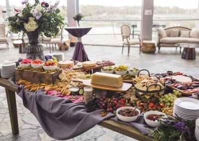Table full of appetizers at a wedding. There is a bunched purple tablecloth and a floral arrangement at the end of the table with plates.