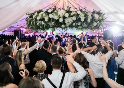 A crowd of wedding guests at the reception dancing with the band on a stage. Overhead there is draped sheer fabric and a large floral arrangement hanging from above.
