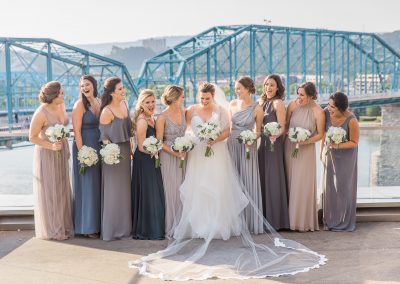A bride in the middle of a row of her bridesmaids on the balcony of the Hunter Museum with the pedestrian bridge in the background. All of the women are holding bouquets and smiling away from the camera.
