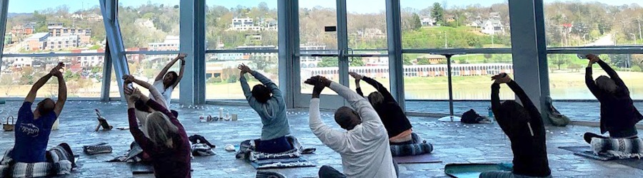 Group of people in the Hunter museum lobby seated on yoga mats stretching to the side with their arms above their heads. An instructor sits at the front of the room.