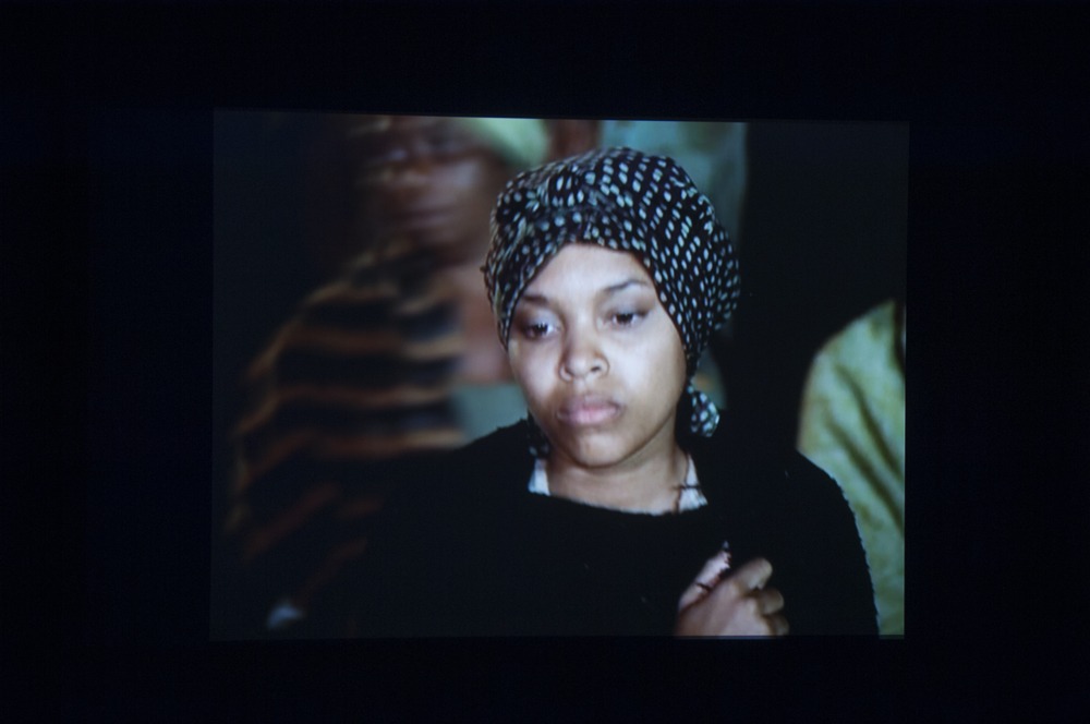 Snapshot from a video on a screen of a woman wearing a head wrap and black shawl looking downwards.