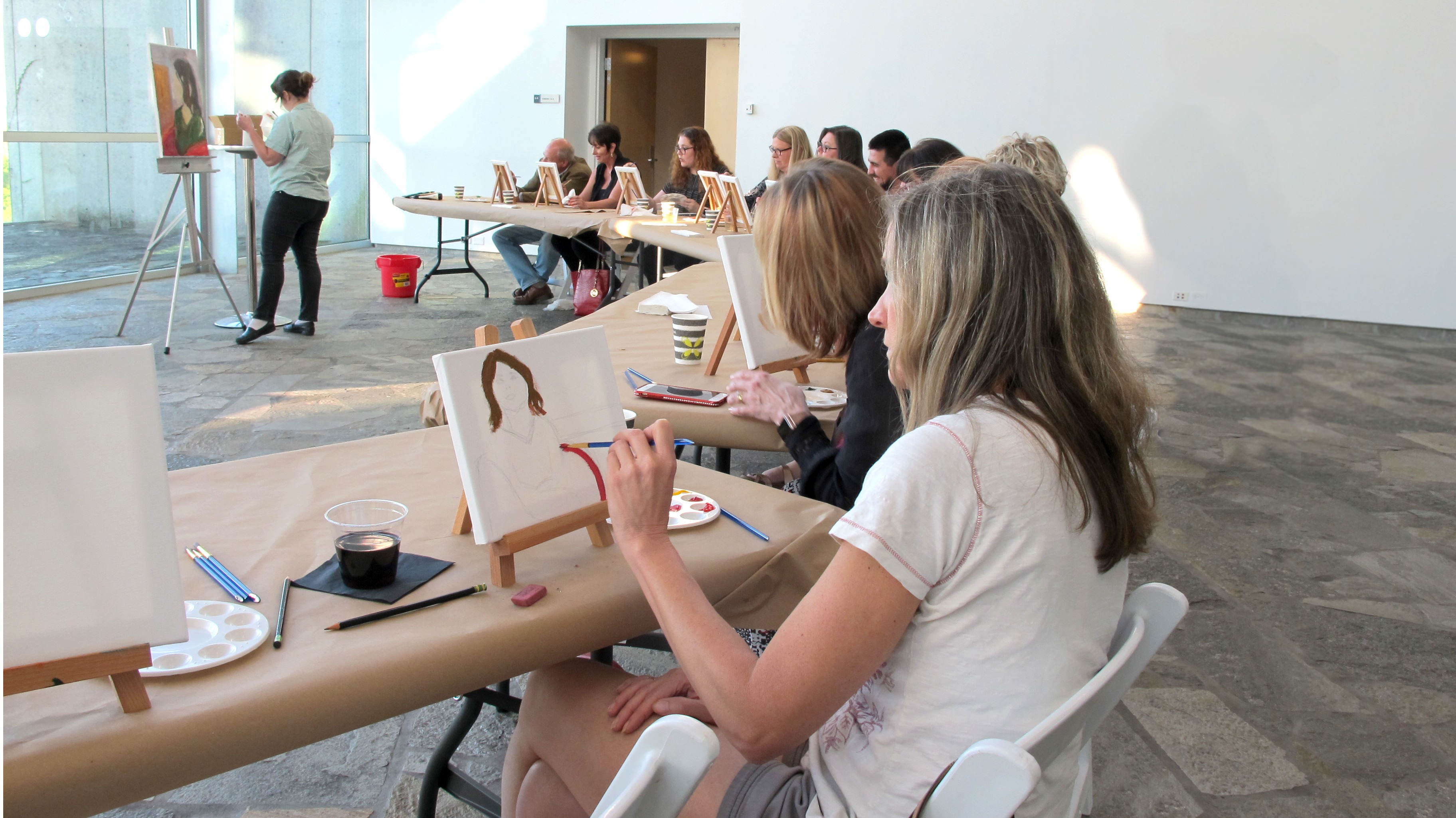 Woman sits at a long table and paints on a small canvas. Other painters sit at tables behind her.