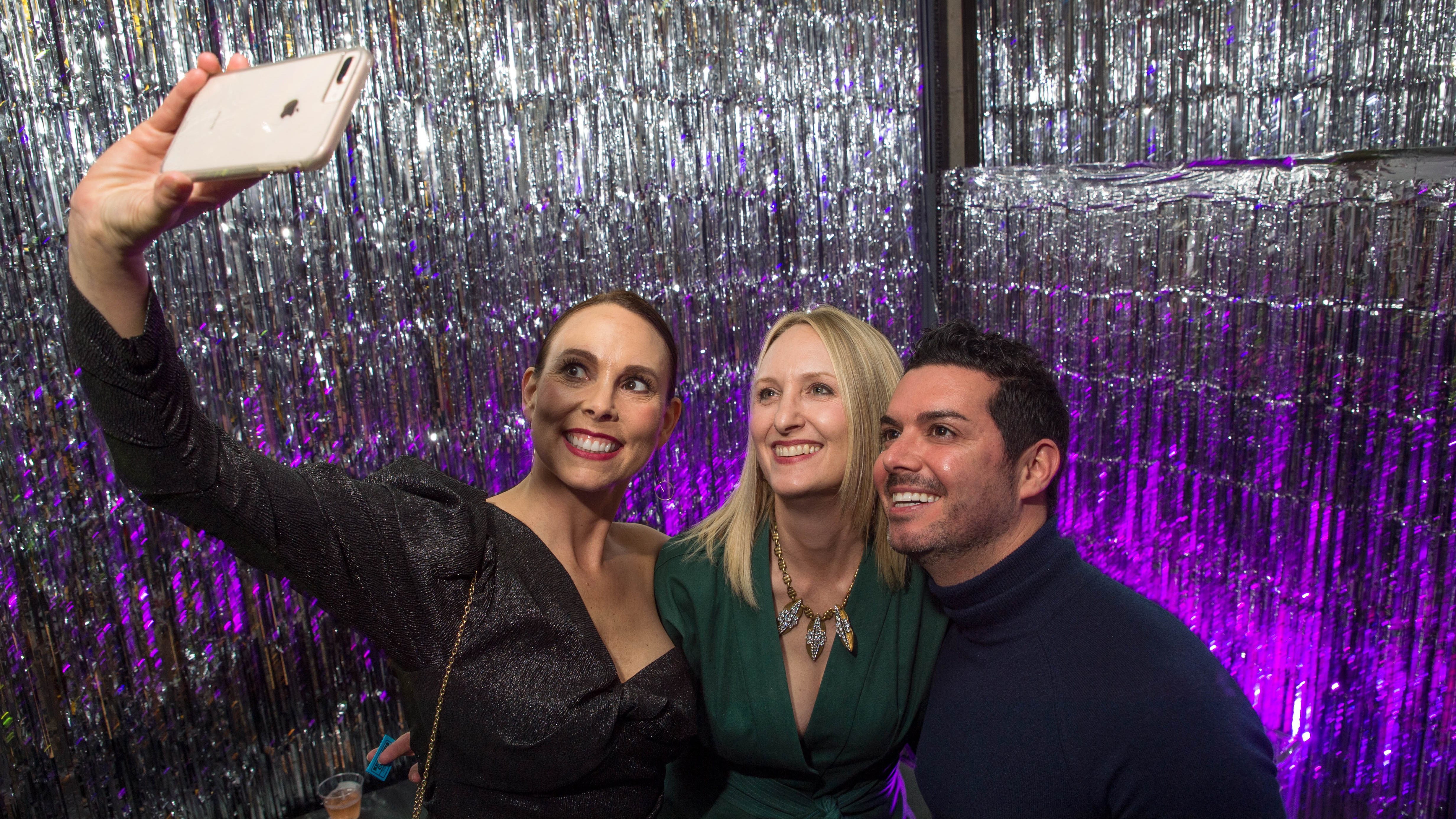 Two women and a man take a selfie against a shimmery silver background with purple lighting.
