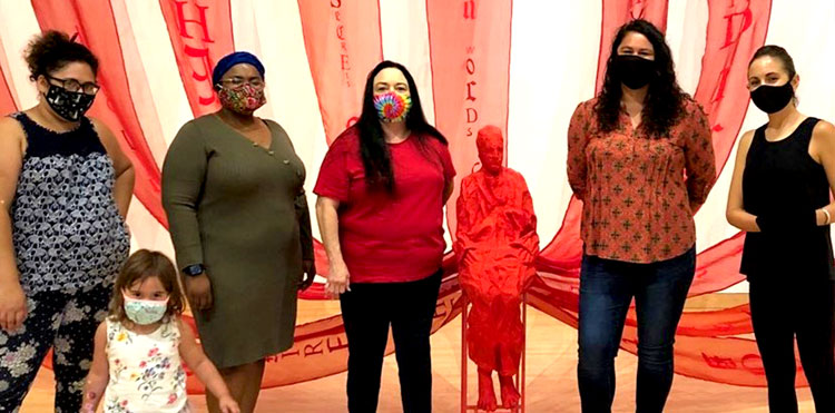Group of five poets and a child standing in front of exhibit which has large flowing pieces of red fabric on the wall that convene at a sculpture of a person cloaked in red sitting in a chair.