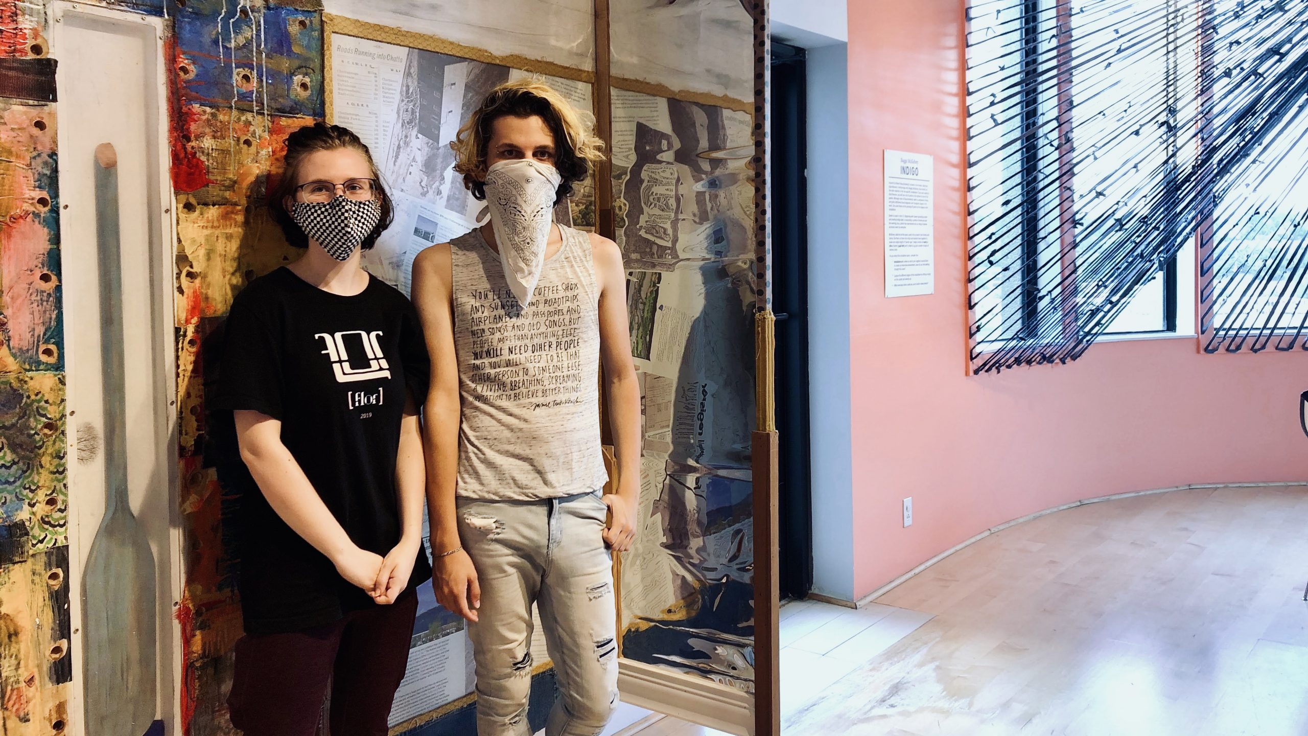 Two teens stand in front of colorful collage on the wall. They are wearing face masks.
